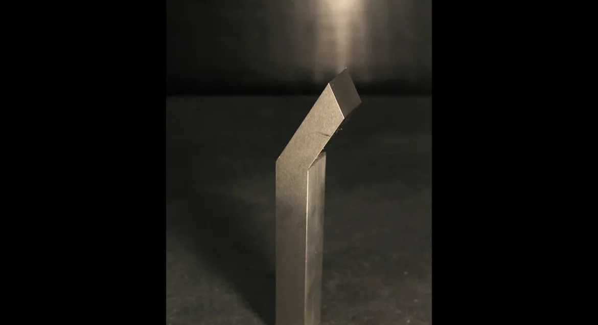 A single tungsten bucking bar under a direct overhead spotlight on a dark bench, lit like a piece under inspection to diagnose a weight problem