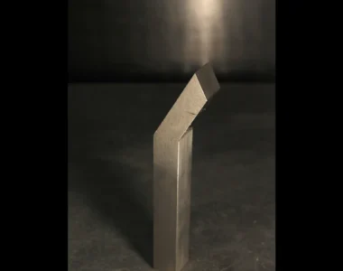 A single tungsten bucking bar under a direct overhead spotlight on a dark bench, lit like a piece under inspection to diagnose a weight problem
