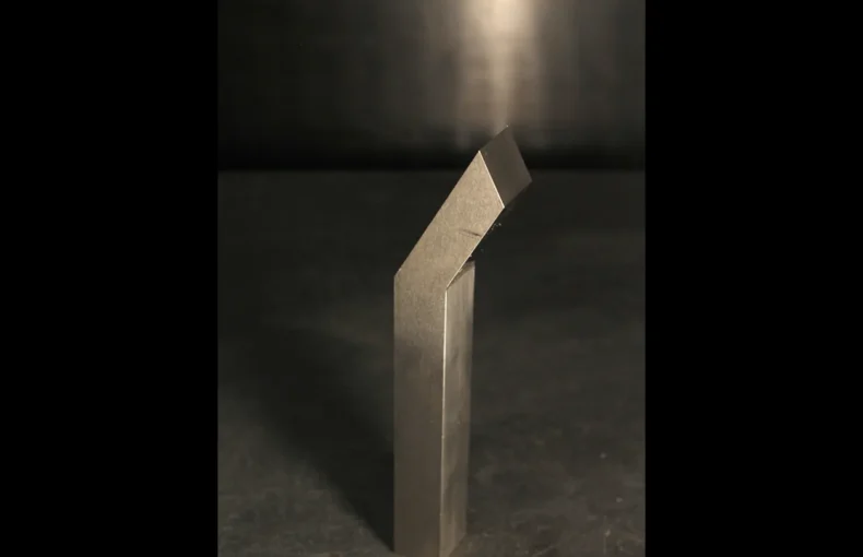A single tungsten bucking bar under a direct overhead spotlight on a dark bench, lit like a piece under inspection to diagnose a weight problem