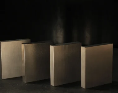 Four flat rectangular tungsten bucking bars standing in a staggered row on a dark bench, the range of weights a rivet gun and bar get matched across