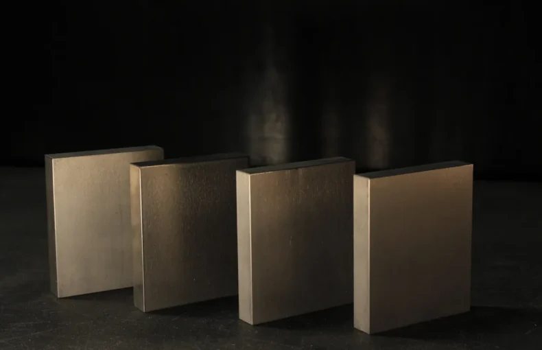 Four flat rectangular tungsten bucking bars standing in a staggered row on a dark bench, the range of weights a rivet gun and bar get matched across